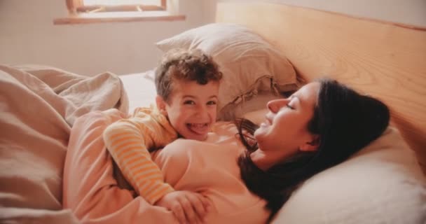 Мальчик лежит в постели с матерью и делает смешное лицо — стоковое видео
