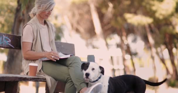 Счастливая пожилая женщина с собакой читает книгу в парке — стоковое видео