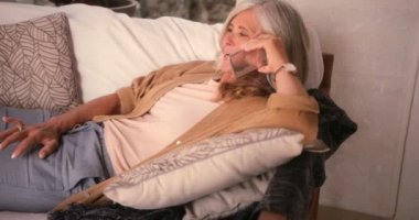 Olgun bir kadın cep telefonuyla konuşuyor ve kanepede dinleniyor.