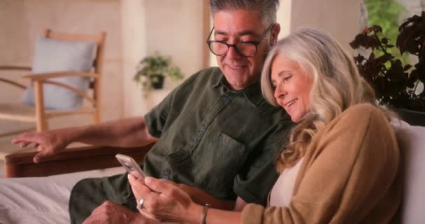 Lykkelig modent par som bruker smarttelefon og slapper av sammen hjemme – stockvideo