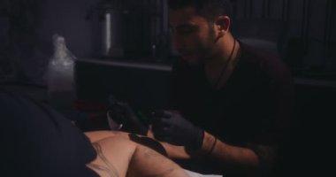 Genç hippi dövme sanatçısı adamın bacağına dövme yapıyor.