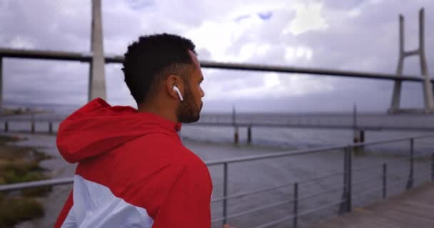 Widok z tyłu afrykańsko-amerykańskiego człowieka ze słuchawkami biegnącymi w pobliżu mostu rzecznego — Wideo stockowe