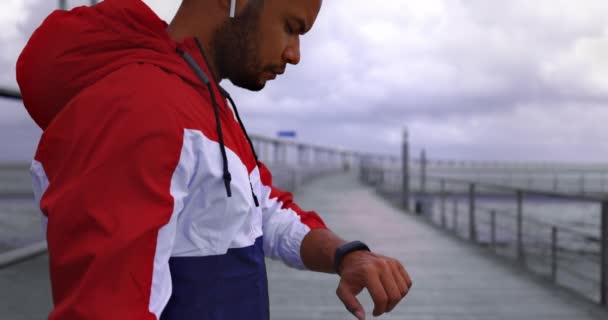 Hombre afroamericano tocando el rastreador de fitness después de correr por el puente del río — Vídeos de Stock