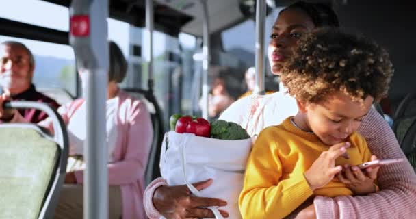Afroamerikanische Mutter hält Lebensmittel in der Hand, Sohn spielt im Bus mit Smartphone — Stockvideo