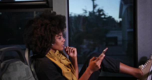 アフリカ系アメリカ人の女性で、イヤホンやスマートフォンが公共バスに座っている — ストック動画