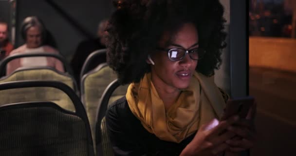 Mulher afro-americana com fones de ouvido mensagens no smartphone sentado no ônibus — Vídeo de Stock