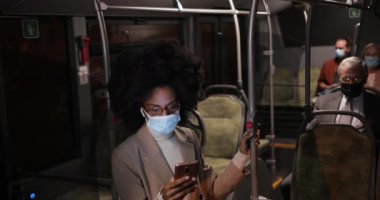 Afro-Amerikalı iş kadını, mavi maske takıyor ve otobüste akıllı telefon tutuyor.