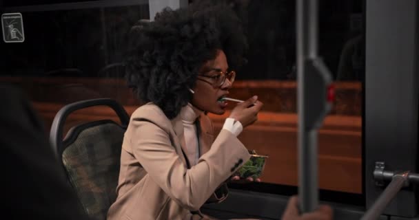 African-american business woman with earphones eating salad on public bus — Stock Video