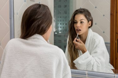 Arkadan görünüşü esmer banyoda ayakta ve sensually yansıma seyir ruj uygulama beyaz bornoz