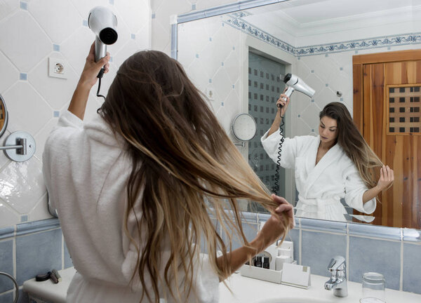 Back view of girl in white bathrobe using hairdryer and styling long hair in modern bathroom
