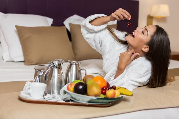 Young attractive woman lying on white and brown bedsheet and eating cherry with plate of different colorful fruits and coffee set put near