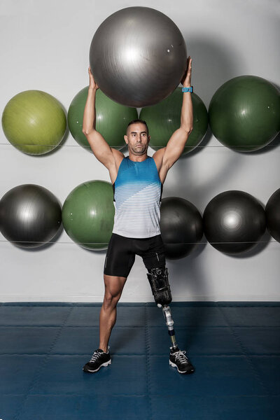 Adult confident sportsman with leg prosthesis holding medicine ball above head training in gym