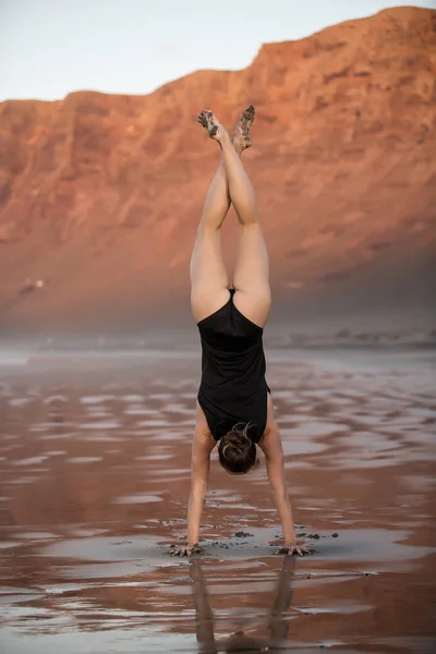Back view of fit woman showing handstand while doing yoga on beach standing on wet sand