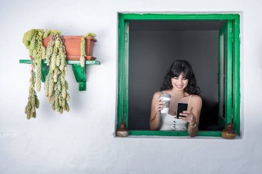 Mutlu genç İspanyol kadın banliyö evinin açık penceresinden dışarıyı gözetlerken sıcak içecek ve akıllı telefona göz atıyor.