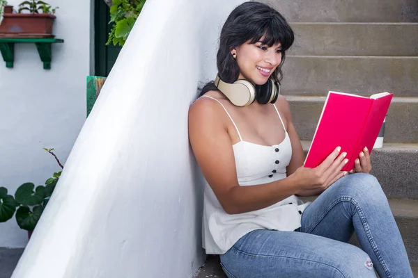 快乐的西班牙女人 戴着耳机 边看有趣的书 边在楼梯上冷着身子 — 图库照片