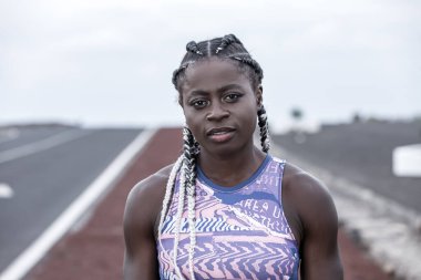 Örgü ören Afro-Amerikalı kadın koşucu açık hava eğitimi sırasında yolun bulanık arka planında dikilirken kameraya bakıyor.