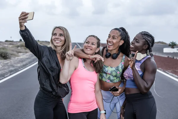 Bir grup mutlu çoklu etnik kadın sporcu yolda durup kenar mahallelerdeki koşu eğitiminde selfie çekerken gülümsüyor ve V işareti yapıyor.