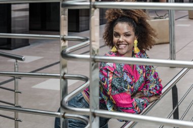 İyimser genç Afro-Amerikan kadın kıvırcık saç stili canlı renkli bluz ve metal korkuluklara yaslanmış moda püsküllü kot pantolon giyiyor ve kameraya bakıyor.