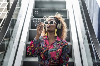 Sarı küpeli, güneş gözlüklü, havalı siyahi bir kadın. Gün ışığında kablosuz kulaklıkla yürüyen merdivenlerde dudaklarını bükerek otururken köpükler saçıyor.