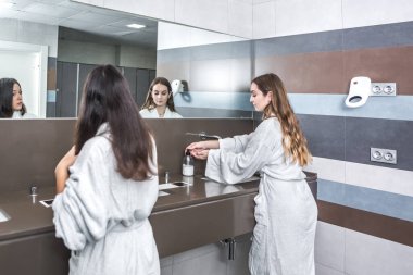 Bornozlu genç bir kadının arka görünüşü lavabonun üstünde ellerini yıkaması kız arkadaşının yanında saçlarını taraması modern spa salonunun tuvaletinde.