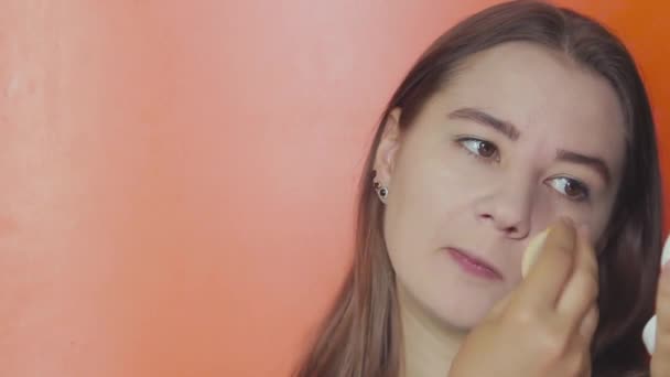 Een jong meisje op een oranje achtergrond brengt stichting op haar gezicht — Stockvideo