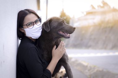 Maskeli ve gözlüklü bir kız salgın sırasında köpeğe sarılıyor.