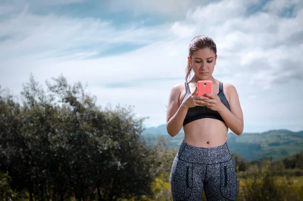 Latino woman with exercise clothes using a cell phone in the middle of nature