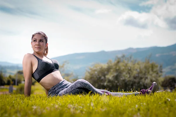 Genç Latin kadın çimenlerin üzerinde rahat egzersiz kıyafetleriyle oturuyor ve sol tarafa bakıyor.