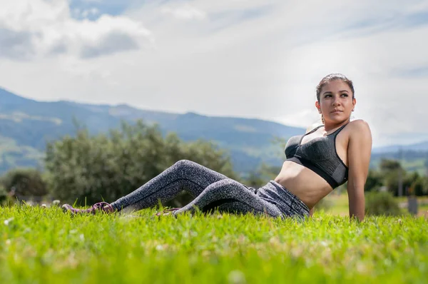 young Latina woman sitting on the grass in relaxed exercise clothes looking at the right side