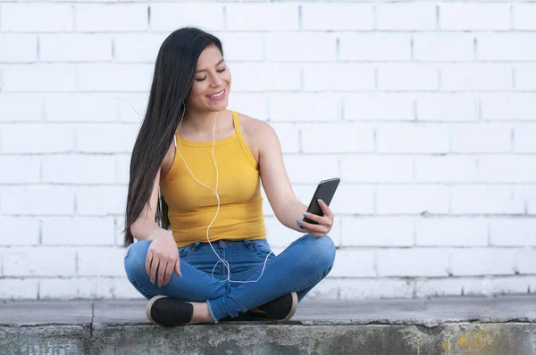 young hispanic woman sitting on a sidewalk texting on her cell phone with copy-space