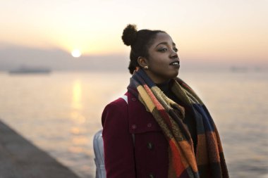 Bir kırmızı ceket ve atkı bir gün batımı ile sular kenar uzaklığı 25 yaşında Afrikalı kadın portresi.