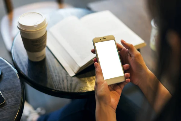 Mobil phone in the womans hand, there is a take away coffee, abook and an empty plastic cup in the scene.