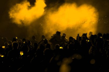 Konser alanı, kalabalık insan ve duman ile sarı sahne ışıkları.