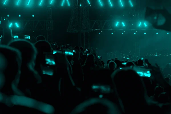 Concert field, crowded people and turquoise stage lights with smoke.