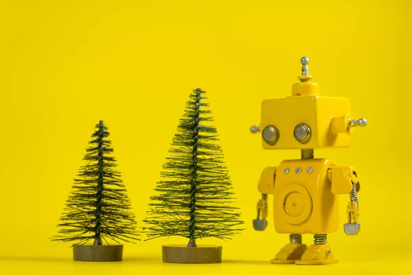 Cute, yellow, handmade robot on a yellow background with two pine trees.