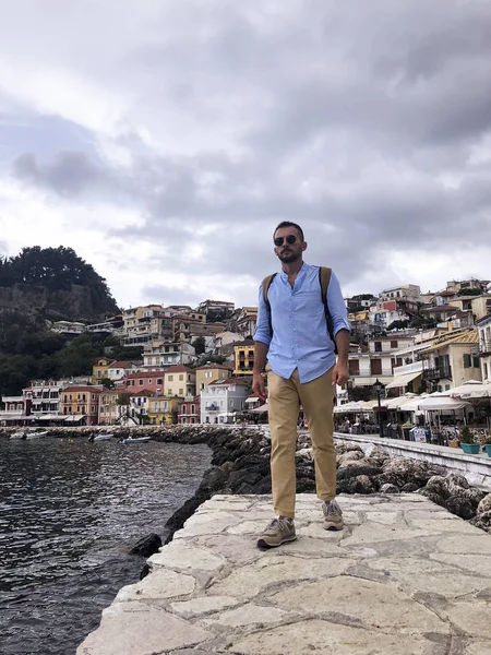 Portrait of a young man with brown pants, blue shirt, sun glasses and a brown backpack at Parga Coast Greece.