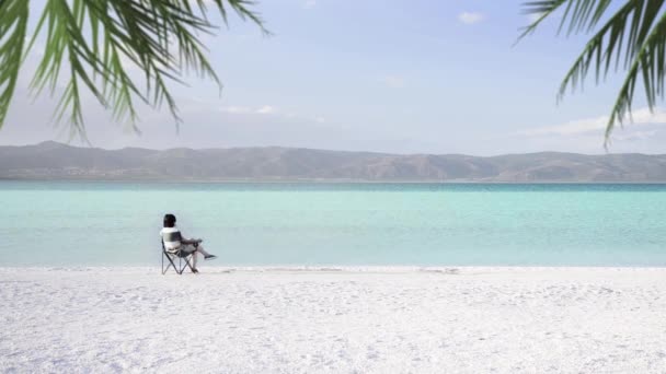 白い砂浜とターコイズ色の水とモルディブのような Salda 水域の端の近くの携帯用の椅子に座っている一人の女性 我々は フレームの隅にいくつかのヤシの葉を参照してください ブルドゥル トルコ — ストック動画