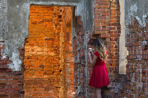 漂亮的女孩穿着红衣服站在被毁的砖墙中间手里拿着智能手机 — 图库照片