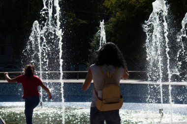 Ukrayna Zhytomir. 14 Eylül. 2020, şehirde sıcak bir günde fıskiyenin arka planında annesi olan kız.