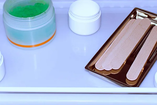 Küçük kavanozlarda kozmetik jel ve bulanık beyaz masa arkaplanındaki tıbbi tepside ahşap spatulalar.