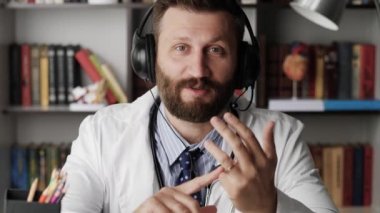 Doktor kamerayla konuşuyor. Beyaz önlüklü çekici sakallı pratisyen doktor kameraya bakarken bir şeyi açıklıyor.