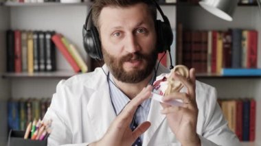Otolaryngologist kameraya konuşuyor. Beyaz önlüklü çekici sakallı kardiyolog insan kulağı modelini kullanarak kameraya bakarken bir şeyi açıklıyor.