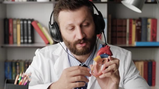 Il medico cardiologo sta parlando con la telecamera. cardiologo barbuto in camice bianco spiega qualcosa guardando la macchina fotografica, nelle sue mani tiene modello di cuore umano e spiega con il suo esempio — Video Stock