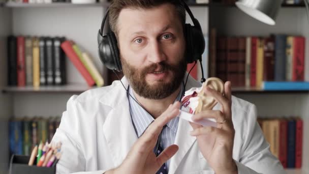 Отоларинголог разговаривает с камерой. Привлекательный бородатый кардиолог в белом халате кое-что объясняет, глядя в камеру, используя в качестве примера модель человеческого уха — стоковое видео