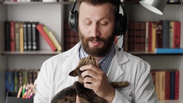 Docteur vétérinaire parlant à la caméra. attrayant barbu médecin vétérinaire en manteau blanc tient chat dans ses mains et explique quelque chose tout en regardant la caméra — Video