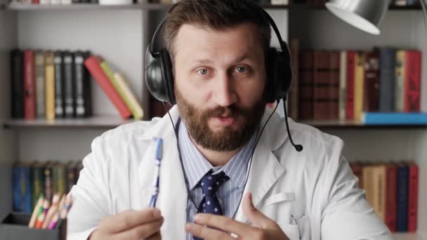 Médico dentista hablando con la cámara. Atractivo médico dentista barbudo sostiene el cepillo de dientes en su mano y habla mientras mira la cámara explicando algo — Vídeos de Stock