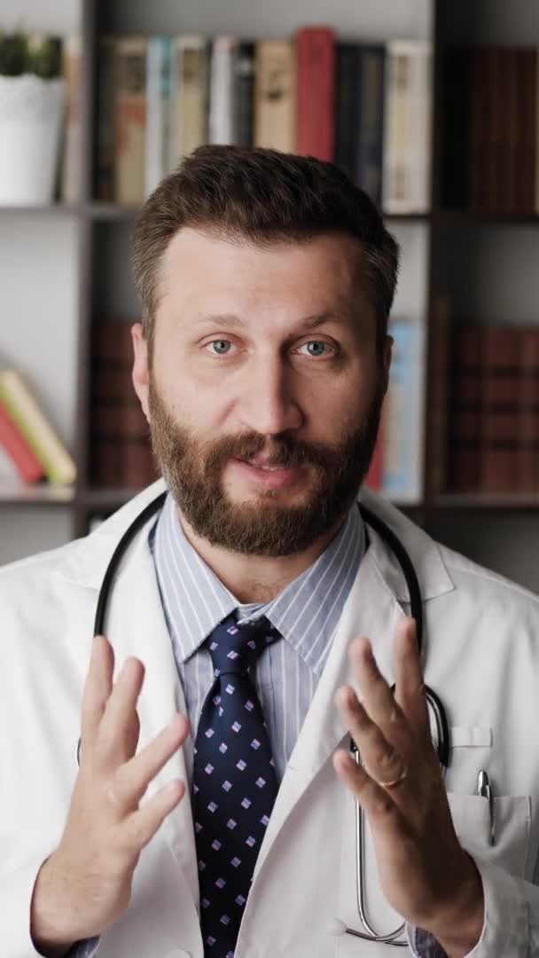 Доктор розмовляє з камерою. Вертикальна орієнтація привабливий бородатий лікар загальної практики в білому пальто пояснює щось, дивлячись на камеру — стокове відео