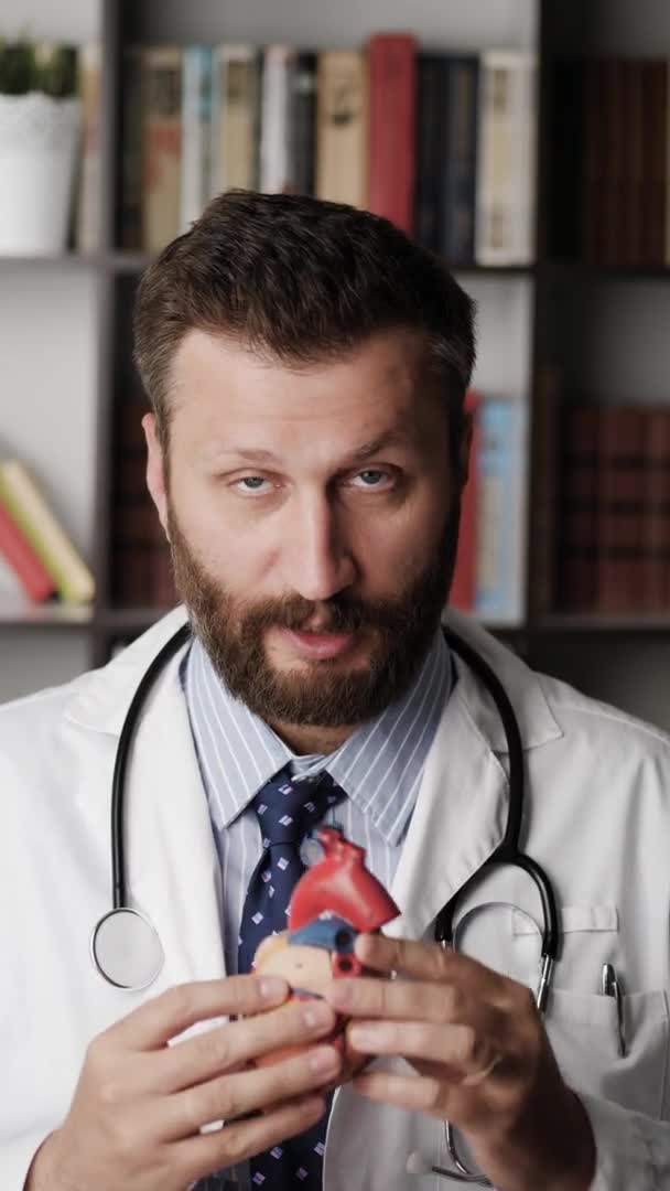 Лікар кардіолог розмовляє з камерою. Вертикальна орієнтація бородатого кардіолога пояснює щось, дивлячись на камеру, в руках він тримає модель людського серця і пояснює своїм прикладом — стокове відео