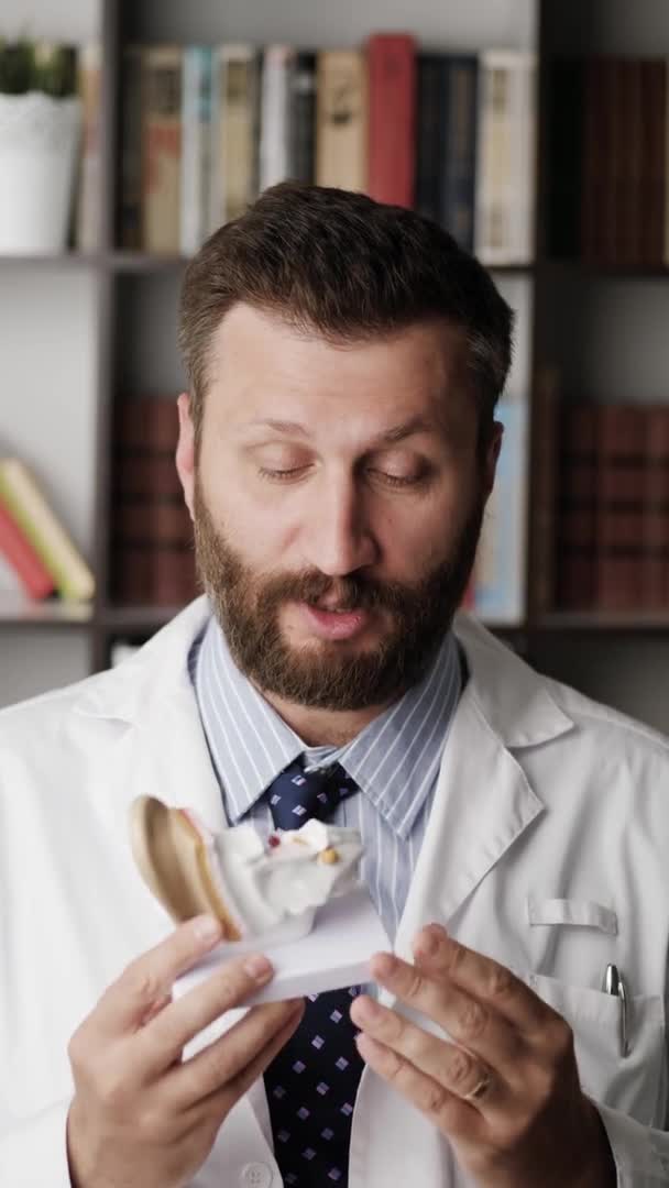 Otolaryngologist talking to camera. Vertical orientation attractive bearded cardiologist in white coat explains something while looking at camera using model of human ear as example — Stock Video