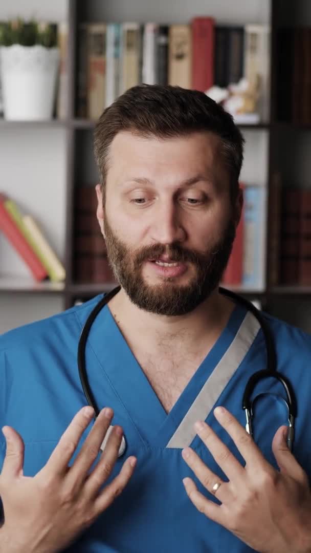 Doktor chirurg mluví s kamerou. Vertikální orientace atraktivní vousatý lékař chirurg drží v ruce falešné umělé srdce a říká něco, zatímco se dívá do kamery — Stock video